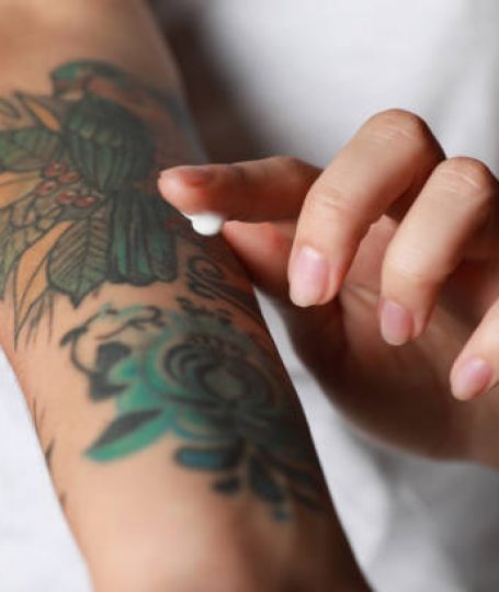 Woman applying cream on her arm with tattoos against black background, closeup